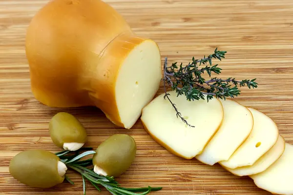Scamorza, zur Hälfte in Scheiben geschnitten; daneben grüne Oliven und Kräuter