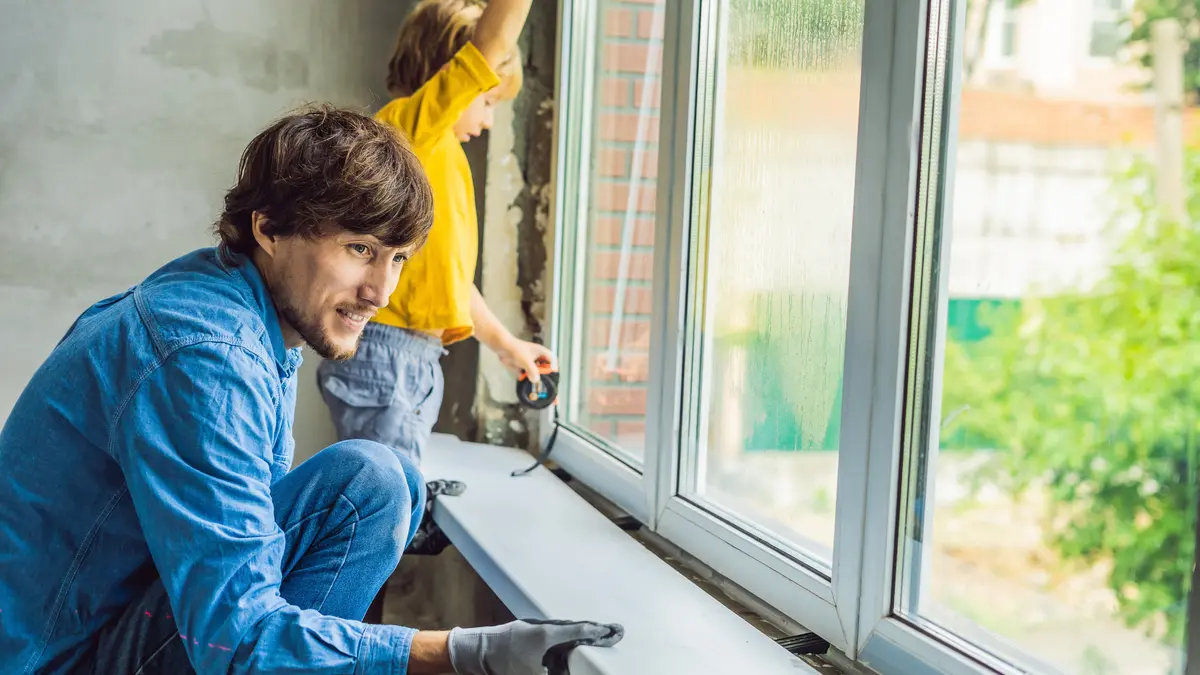 Ein Mann tauscht ein Fenster aus. Er montiert gerade die Fensterbank an oder ab. Ein kleiner Junge unterstützt ihn.