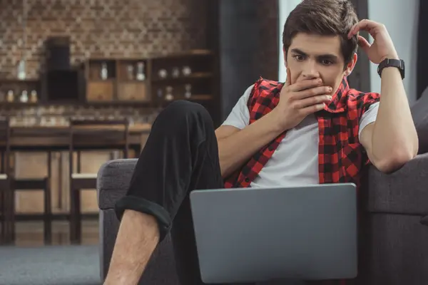 junger Mann auf dem Sofa mit Laptop blickt erschrocken
