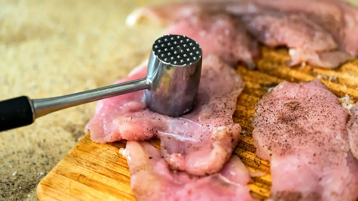 Rohe Schnitzel, mit Fleischzartmacher gewürzt, liegen auf einem Brett. Ein Fleischklopfer ruht auf einem Schnitzel.