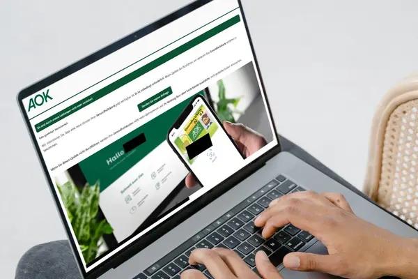 Zwei Hände auf der Tastatur eines aufgeklappten Laptops, auf dem Display eine Phishing-Mail mit AOK-Bezug