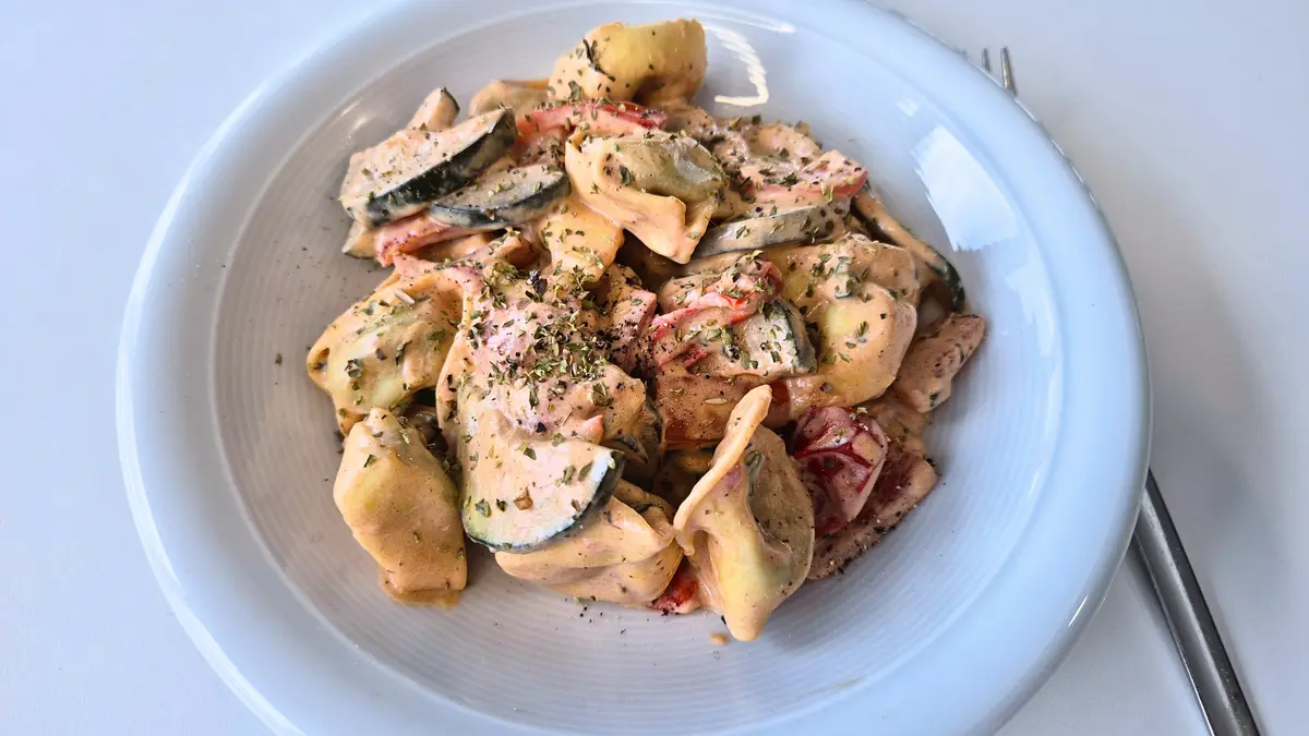 Eine weiße, tief geschwungene Schale mit Tortellini in cremiger Soße, garniert mit Kräutern und kleinen Gemüse- und Tomatenstücken.