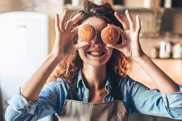 Eine junge Frau mit Kochschürze steht lachend in einer Küche und hält sich zwei Muffins vor die Augen 