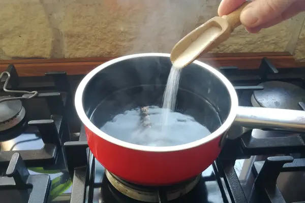 Ein roter Topf mit heißem Wasser steht auf einem Gasherd. Eine Hand gibt mit einem Holzlöffel Salz in das Kochwasser.