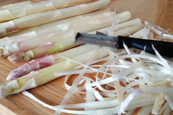 Mehrere Stangen geschälter, weißer Spargel liegen zusammen mit einem Spargelschäler und Spargelschalen auf einer Arbeitsplatte aus Holz.