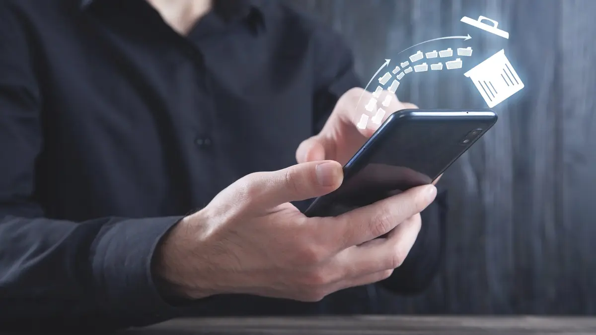 Ein Mann in einem schwarzen Hemd hält ein Smartphone in den Händen. Über dem Display schwebt eine digitale Illustration, die zeigt, wie mehrere Aktenordner-Symbole in einen Papierkorb fliegen, was den Vorgang des Löschens oder Aufräumens von Daten symbolisiert.