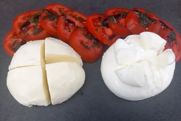 Auf einer schwarzen Arbeitsplatte liegen je ein Stück Mozzarella und ein Stück Burrata, darüber in Scheiben aufgeschnittene Tomaten.