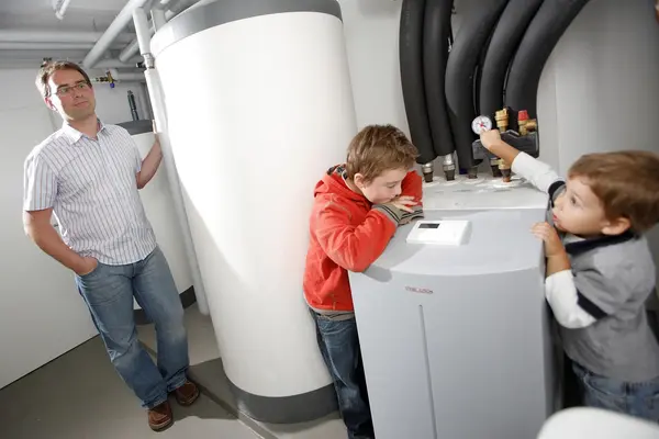 Ein Vater steht im Keller seines Hauses neben seiner Wärmepumpe. Zwei Kinder sind auch anwesend.