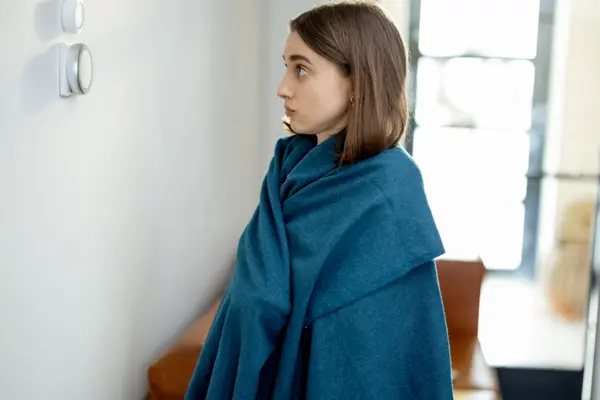 junge Frau, in eine blaue Decke gehüllt, blickt ernst auf einen Heizungsregler.