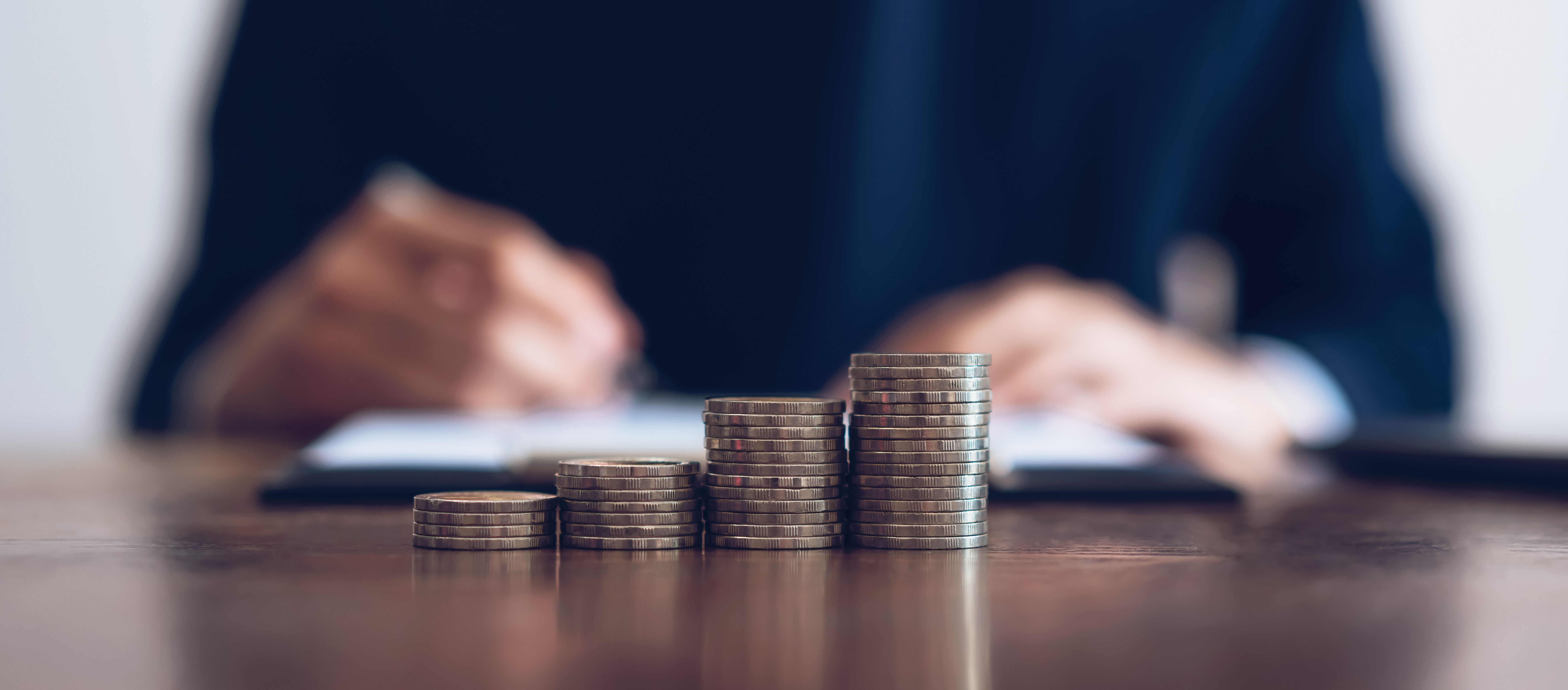 Eine Person notiert sich etwas hinter vier verschieden hohen Stapeln von Geldmünzen.