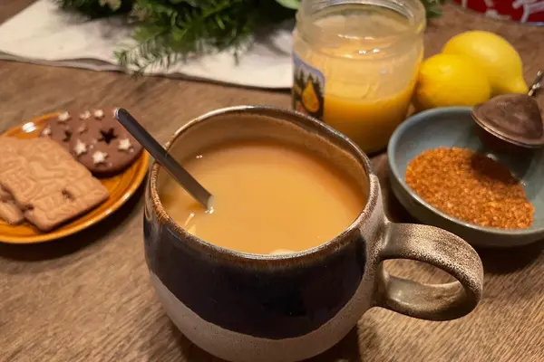 Eine Tasse hellbrauner Tee mit Löffel steht auf einem Holztisch, umgeben von Honig, Zitronen, losen Teeblättern und einem Teller mit Weihnachtsgebäck.