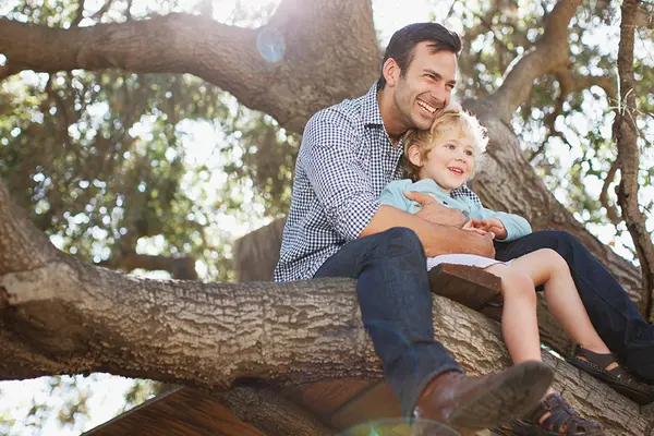 Vater und Sohn sitzen lachen auf einem Baum, der Vater hält das Kind fest