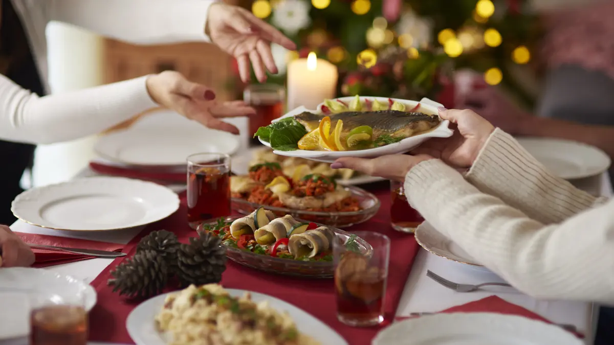 Menschen, die Teller mit Weihnachtskarpfen über festlich gedecktem Tisch passieren, im Hintergrund sieht man einen geschmückten Weihnachtsbaum