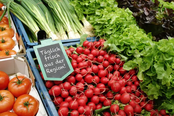 Auf einem Marktstand liegen Gemüse und Salate aus, ein Schild weist auf regionale Hersteller hin.