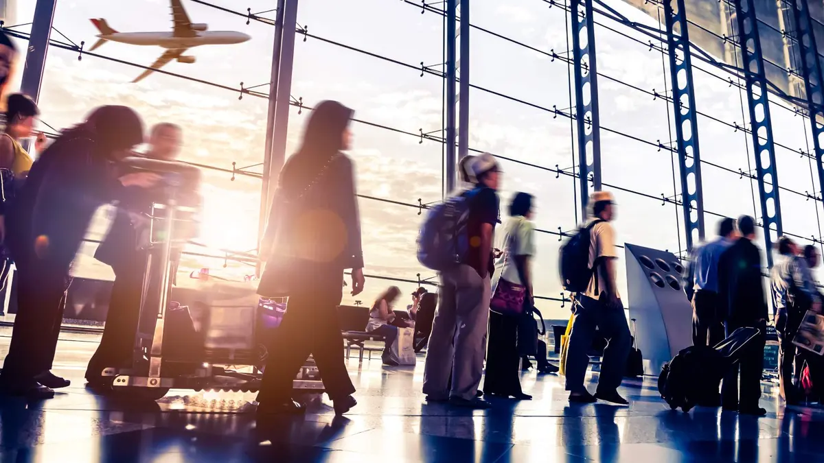 in geschäftiges Flughafenterminal mit Passagieren in Bewegung, die als verschwommene Silhouetten vor einer großen Glasfassade dargestellt sind. Im Hintergrund ist ein Flugzeug beim Start durch das Fenster zu sehen, während helles Sonnenlicht den Raum flutet und lange Schatten sowie Lichtreflexe auf dem glänzenden Boden erzeugt.
