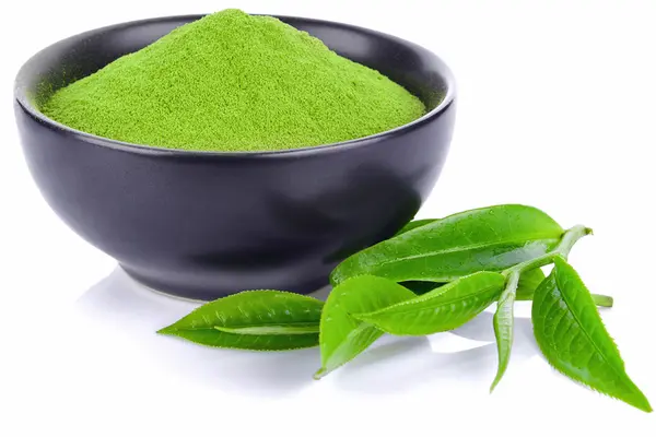 Grünes Matcha-Pulver in einer schwarzen Keramikschale und Teeblätter davor.