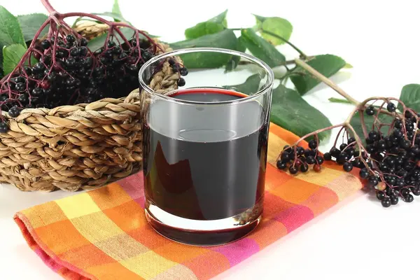 Ein Glas mit Holundersaft steht auf einem bunt kariertem Geschirrtuch, daneben Zweige mit Holunderbeeren.