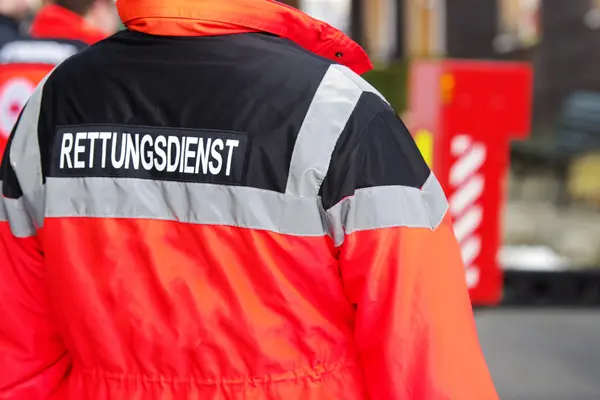 Jemand steht an einer Straße und trägt eine Warnweste, auf der "Rettungsdienst" steht.