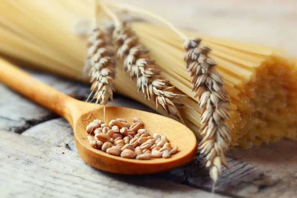 Rohe Spaghetti liegen auf einem Holztisch, darauf eine Weizenähre, daneben ein Holzlöffel mit Weizenkörnern.