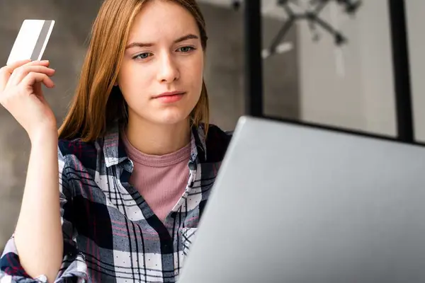 Eine Frau sitzt mit skeptischem Blick vor einem Laptop, sie hält eine Kreditkarte in der Hand