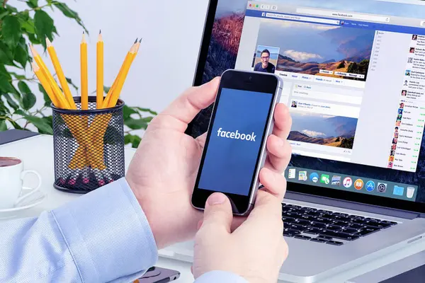 Mann am Schreibtisch hält Smartphone mit dem Schriftzug facebook auf dem Display in seinen Händen, dahinter ein aufgeklappter Laptop mit geöffneter Facebook-Seite
