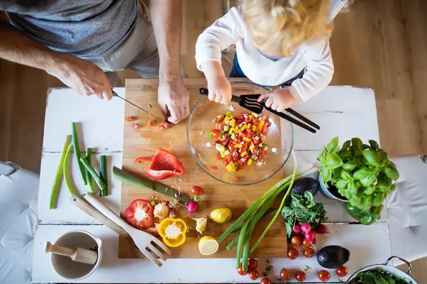 Aus der Vogelperspektive bereiten ein Erwachsener und ein Kind gemeinsam einen bunten Salat mit frischem Gemüse und Kräutern zu. Auf dem Tisch liegen verschiedene Zutaten wie Paprika, Tomaten, Radieschen, Frühlingszwiebeln, Basilikum und Avocado, während in einer Schüssel bereits gemischtes Gemüse liegt.