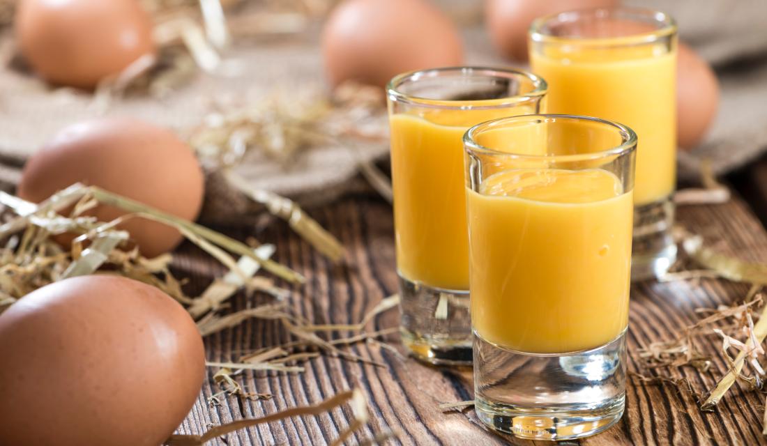 Drei Gläser Eierlikör stehen auf einem Brett, darum liegen Eier und Stroh