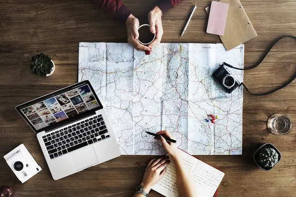 Zwei Personen planen eine Reise an einem Tisch mit Laptop, Weltkarte, Fotoapparat und weiteren Utensilien.