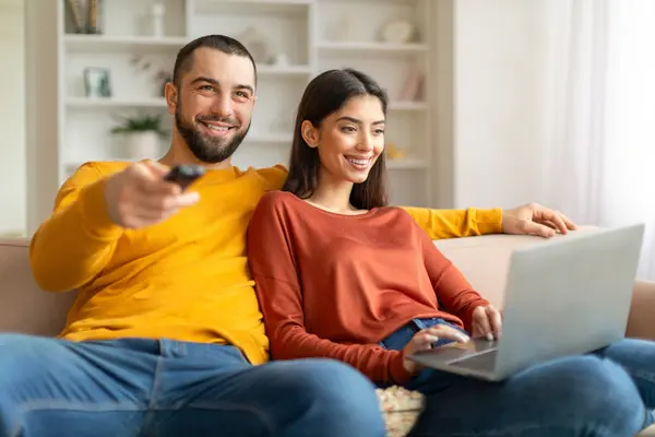 Mann und Frau sitzen auf dem Sofa, Frau hat Laptop auf dem Schoß, Mann hält Fernbedienung in der Hand und schaut TV. Beide lachen.