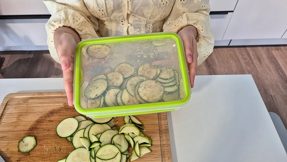 Eine Person in einem cremefarbenen Lochmuster-Oberteil hält eine rechteckige, grüne Plastikdose mit transparentem Deckel in die Kamera. In der Dose sind panierte Zucchinischeiben zu sehen.