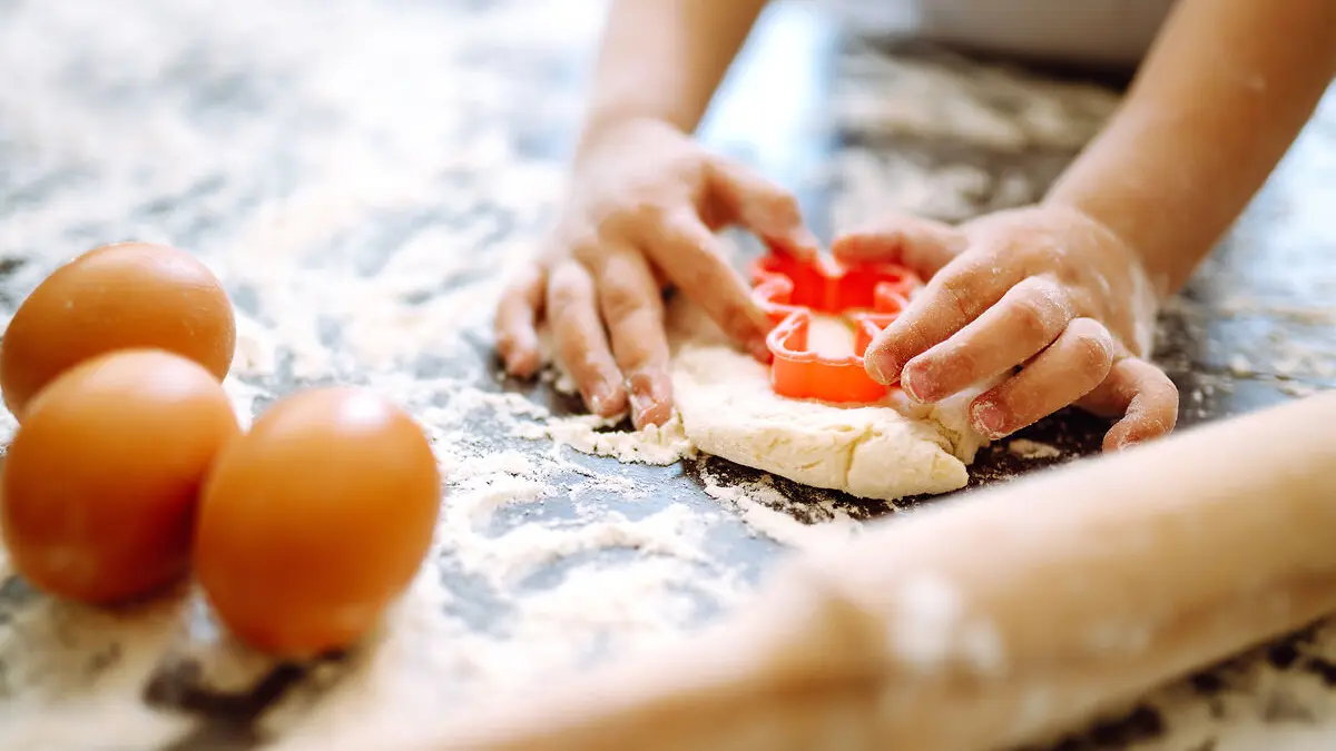 Kinderhände stechen mit einem Teigausstecher eine Form aus einem rohen Teig. Daneben liegen drei rohe Eier und ein Teigroller..