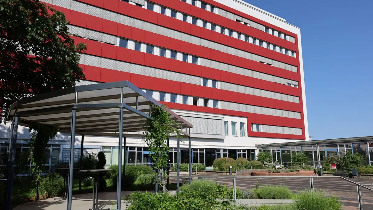 Mehrstöckiges Gebäude mit gebänderter Fassadengestaltung. Davor ein Park mit Sitzmöglichkeiten.