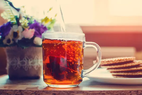 Eine Tasse Tee steht auf einem Tisch, links davon stehen Blumen, rechts davon ein Teller mit Keksen