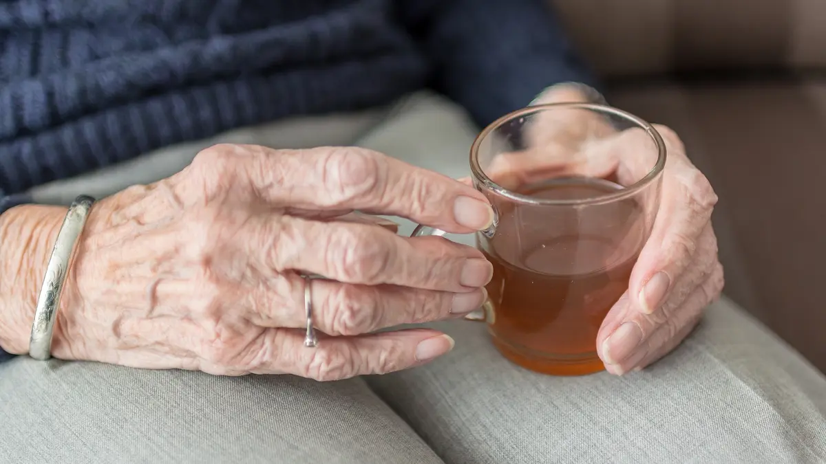 Die Hände einer älteren Dame halten ein Glas mit Schwarztee. Sie stellt das Glas auf ihrem Schoß ab. An ihrer Hand sieht man ein silbernes Armband und einen silbernen Ring mit kleinem weißen Stein. Ihre Hose ist hellgrau, ihr Pullover dunkelblau.