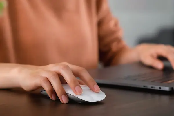 Eine Frau sitzt an ihrem Notebook und hält ihre Hand über eine Computermaus.