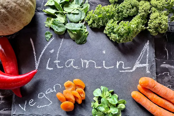 Verschiedene Vitamin A-haltige Gemüse wie Spinat und Möhren liegen auf einer schwarzen Schiefertafel auf der "Vitamin A" und "vegan" steht.