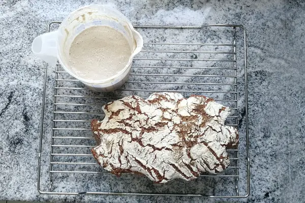 Neben einem Gefäß mit Sauerteig liegt ein fertig gebackenes Sauerteigbrot.