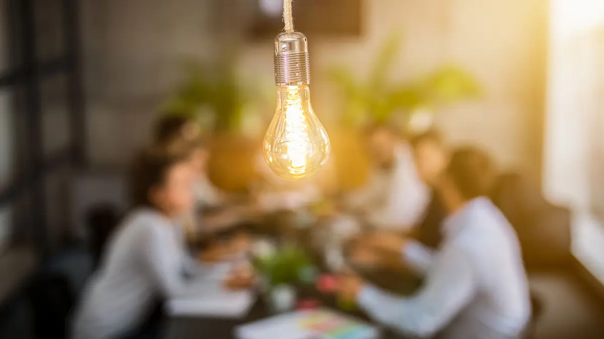 Eine Gruppe Menschen sitzt um einen Tisch und diskutiert, im Vordergrund symbolisiert eine Glühbirne die Ideen