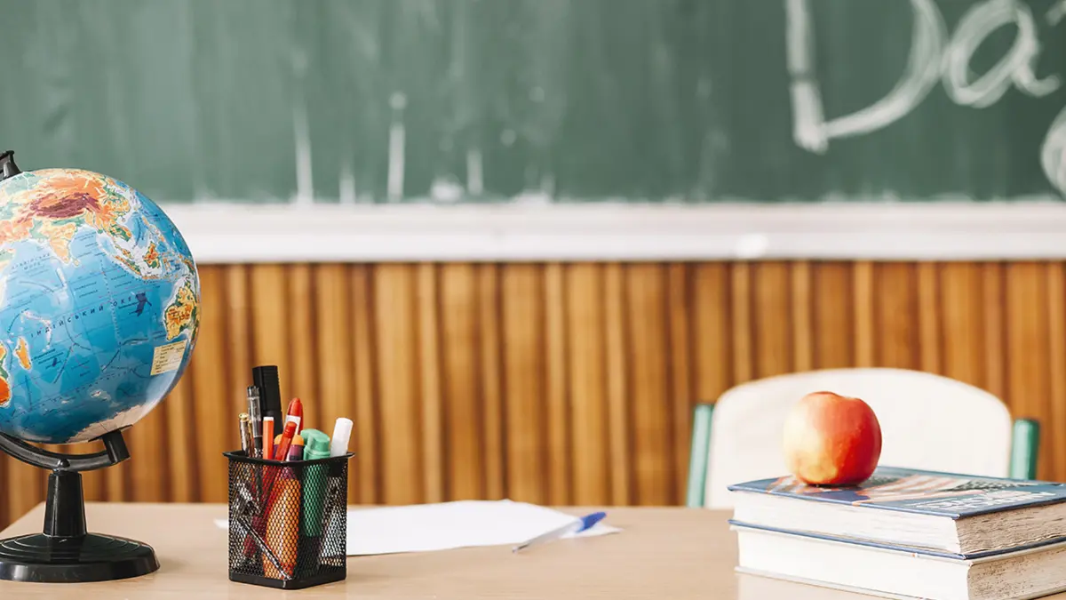 Lehrerpult mit Globus, Stiften, Büchern und einem Apfel vor einer grünen Kreidetafel.