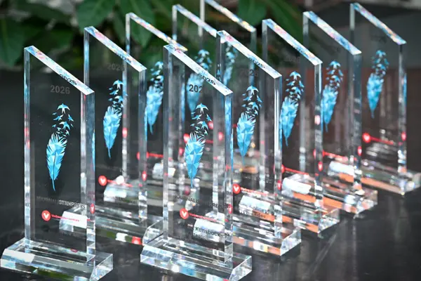 Transparente Trophäen aus Plexiglas stehen aufgereiht auf einem Tisch, jede trägt das Bild einer blauen Feder, aus der am oberen Bereich Vögel aufsteigen.