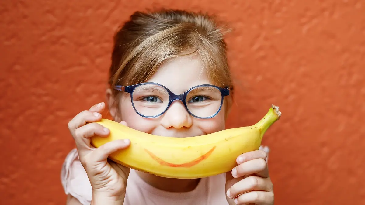 Ein Mädchen mit Brille hält sich einen Banane mit einem aufgemalten Lächeln vor den Mund.