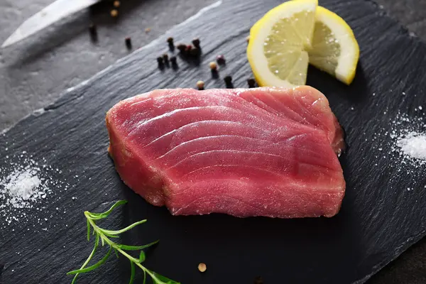 Ein rohes Stück Thunfisch liegt auf einem schwarzen Brett, daneben eine Zitronenscheibe, schwarze Pfefferkörnern und ein Zweig Rosmarin.