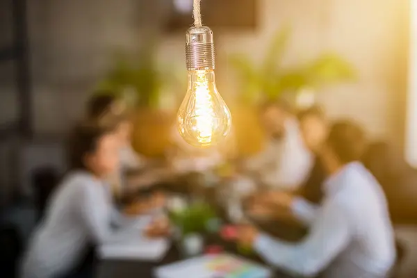Eine Gruppe Menschen sitzt um einen Tisch und diskutiert, im Vordergrund symbolisiert eine Glühbirne die Ideen 