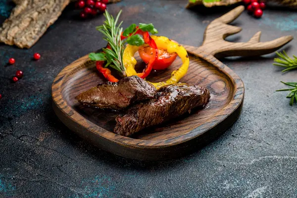 Ein Holzteller in Form eines Geweihs mit einem Stück gebratenem Wildfleisch drauf.