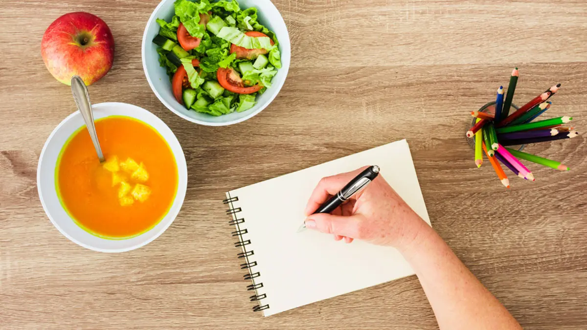Ein Tisch mit Suppe und Salat in Schälchen, einem Apfel und Buntstiften. Eine Hand schreibt auf einen Block