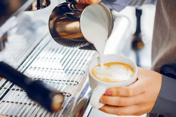 Eine Person gießt aus einer Edelstahlkanne aufgeschäumte Milch in eine Cappuchinotasse