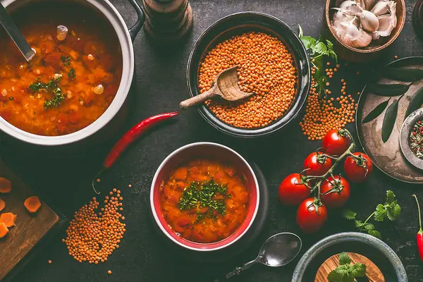 In verschiedenen Töpfen sieht man gekochte und rohe rote Linsen, daneben Zutaten wie Karotten, Tomaten oder Knoblauch
