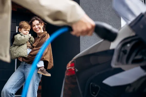 Familie stehen neben einem schwarzen Elektro-Auto, das an einer Ladestation geladen wird.