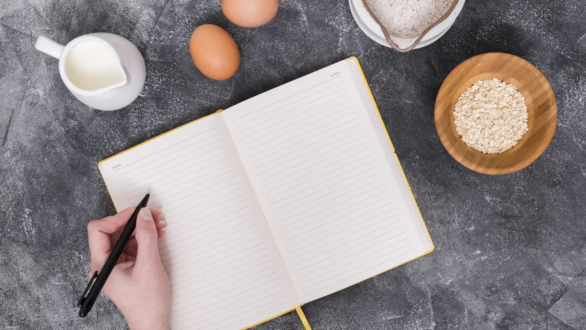 Hand mit Stift und einem leeren Notizbuch, Eier, Milch, Haferflocken an der Seite um das Notizbuch herum.