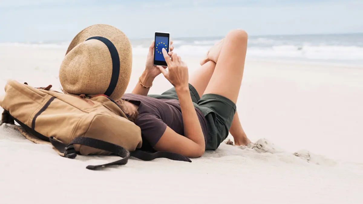 Eine Frau liegt im weißen Sand eines Strandes, Wellen unscharf im Hintergrund. In der Hand hält sie ein Smartphone mit der EU-Flagge auf dem Display.
